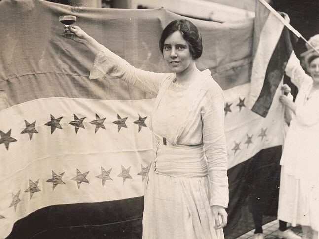 Alice Paul, full-length portrait, standing, facing left, raising glass with right hand